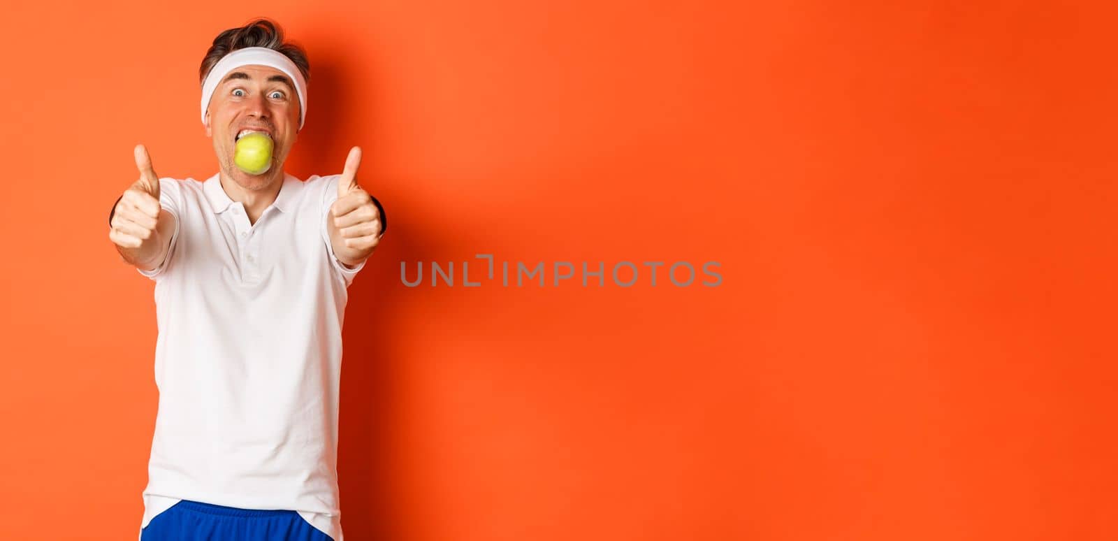 Image of funny middle-aged guy in fitness clothing, holding apple in mouth, showing thumbs-up, standing over orange background.