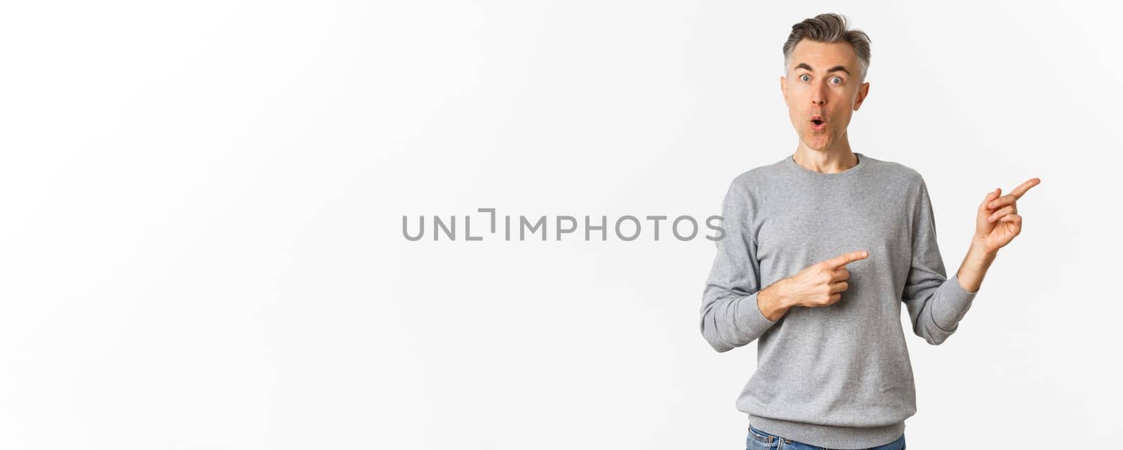 Amazed handsome guy in grey sweater, pointing fingers right and looking curious, askign question about product, standing over white background.