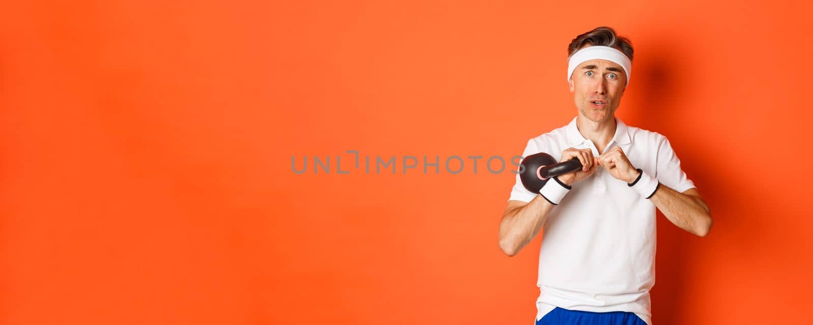 Concept of workout, gym and lifestyle. Close-up of weak and slim middle-aged fitness guy, doing sport exercises with kettlebell, standing over orange background.