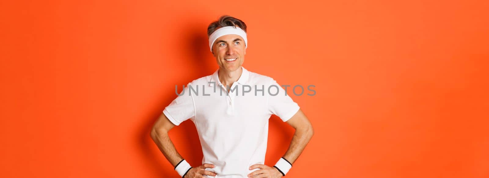 Portrait of satisfied middle-aged man doing sports, looking pleased at upper left corner and smiling, workout to stay healthy, standing over orange background.