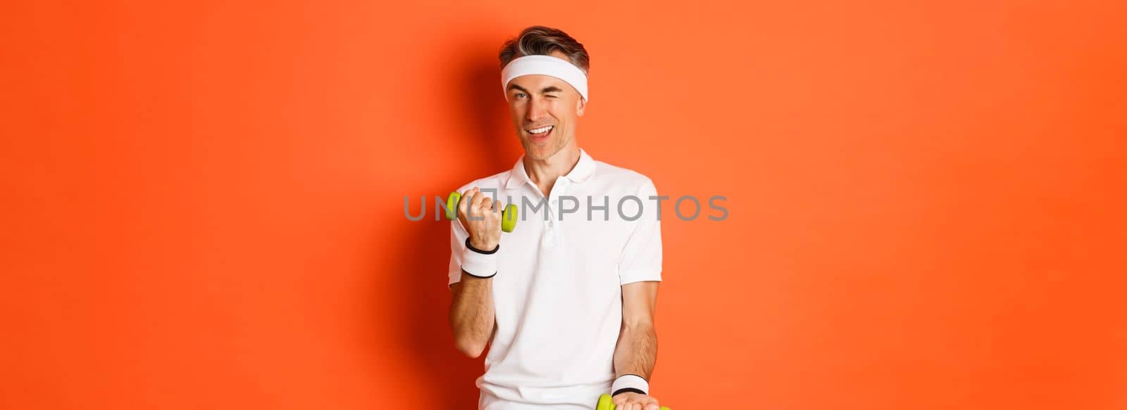 Portrait of handsome cheeky, middle-aged male athlete doing sports, exercise with dumbbells and winking at camera, standing over orange background.