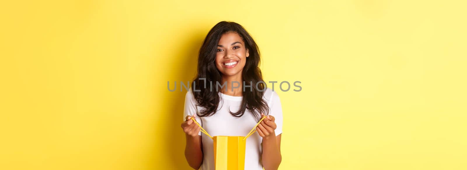 Portrait of happy and delighted african-american girl, open a shopping bad and smiling pleased, thanking for gift on holiday, standing over yellow background.