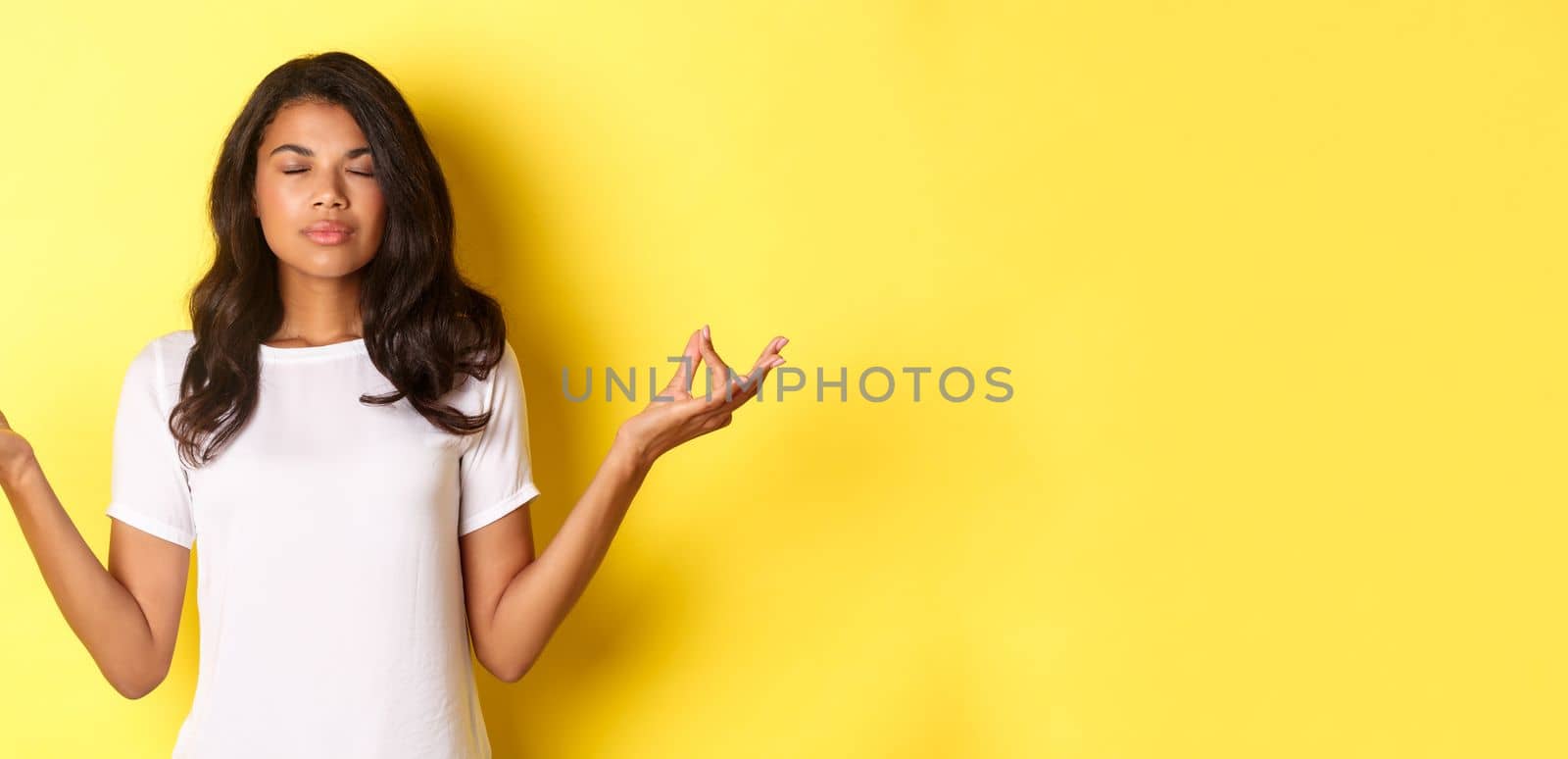 Portrait of relaxed and patient african-american woman, meditating with eyes closed and hands spread sideways, doing yoga over yellow background.