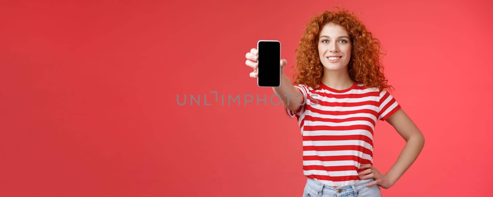 Confident good-looking redhead curly woman present cool app hold smartphone extended hand show phone screen smiling assertive recommending subscribe her blog social-media. Copy space