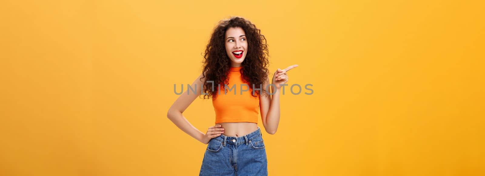 Portrait of delighted enthusiastic stylish young woman. with curly hairstyle in red lipstick and cropped top holding hand on waist pointing and looking left amused and happy over orange background.