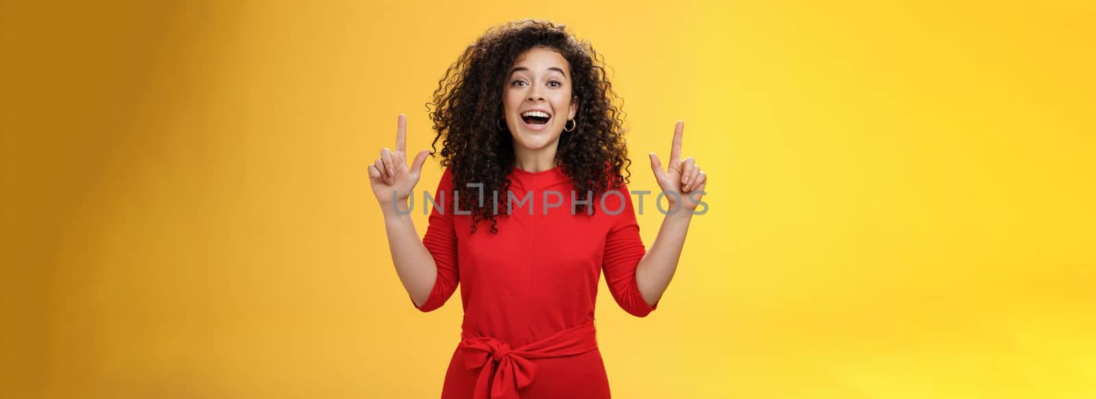 Enthusiastic happy curly-haired young european woman feeling happy present awesome copy space, raising hands pointing up and smiling with delight and admiration over yellow background.