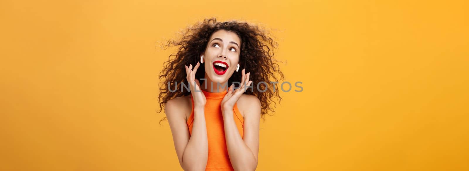 Waist-up shot of attractive silly curly-haired european female in cropped top using wireless earphones touching earbuds and gazing at upper right corner delighted and carefree over orange wall by Benzoix