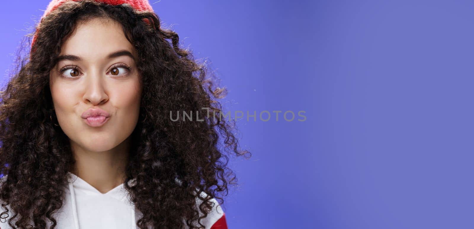 Headshot of funny and playful girl fooling around making faces not scared being funny folding lips like duck and squinting ice, aping standing childish and hilarious over blue wall in winter hat.