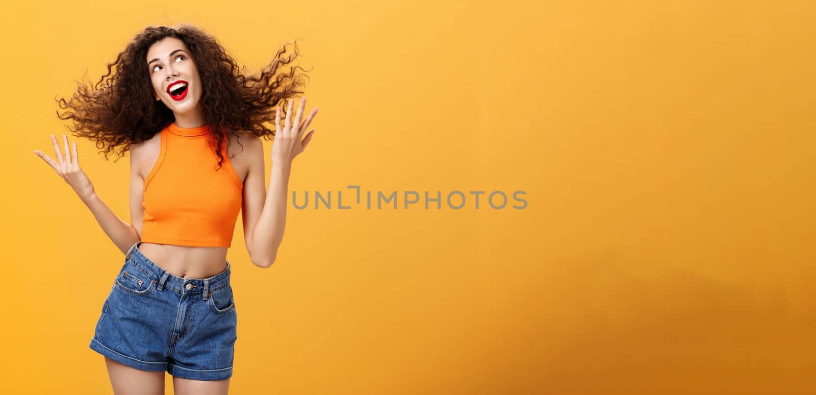 Portrait of silly positive and energized cute female. with curly hairstyle in cropped top flicking hair strand and smiling charming gazing at upper right corner dreamy posing happy over orange wall.