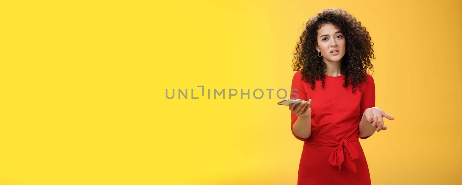 So waht, I confused. Questioned uncertain woman with curly hair in red dress shrugging looking clueless as holding hand cannot understand where order as checking mail box via device over yellow wall. Technology and internet concept