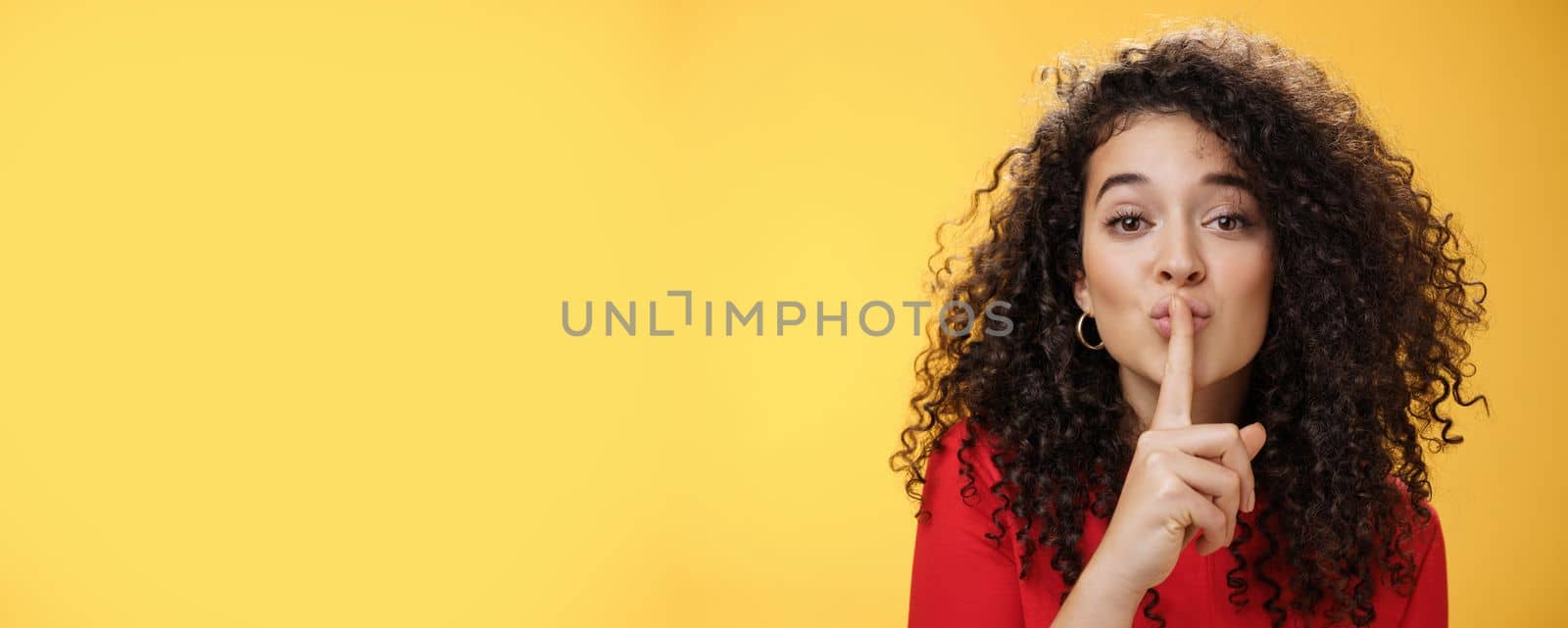 Shh please not tell secret. Cute and tender woman with curly hairstyle gently shushing with index finger on folded lips asking keep promise and remain silent, making surprise over yellow wall by Benzoix