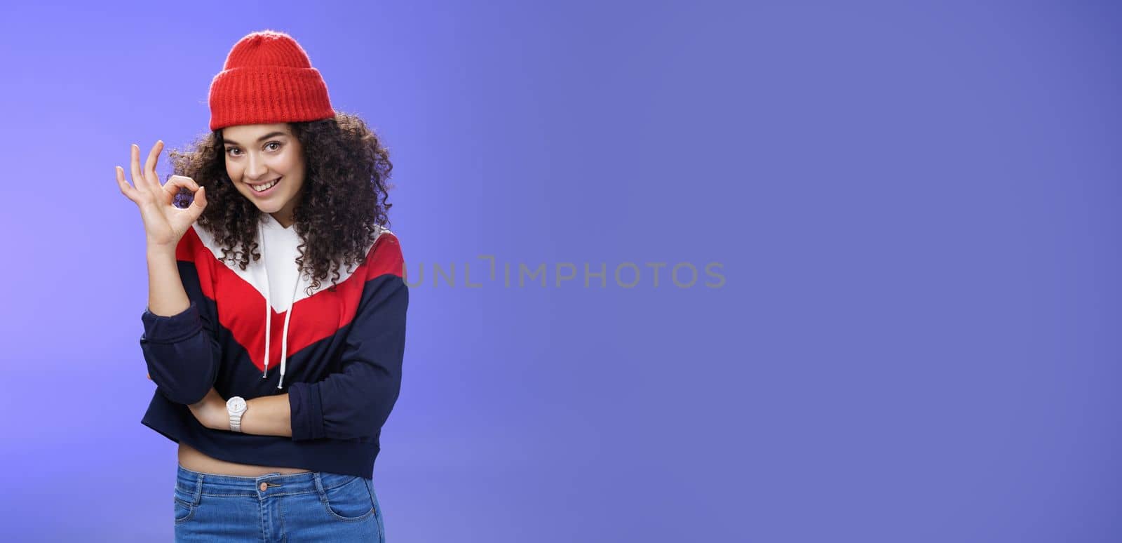 Portrait of pleased good-looking assertive woman with confident look and curly hair wearing warm winter hat showing okay gesture and smiling assuring, liking and approving awesome outfit of friend by Benzoix