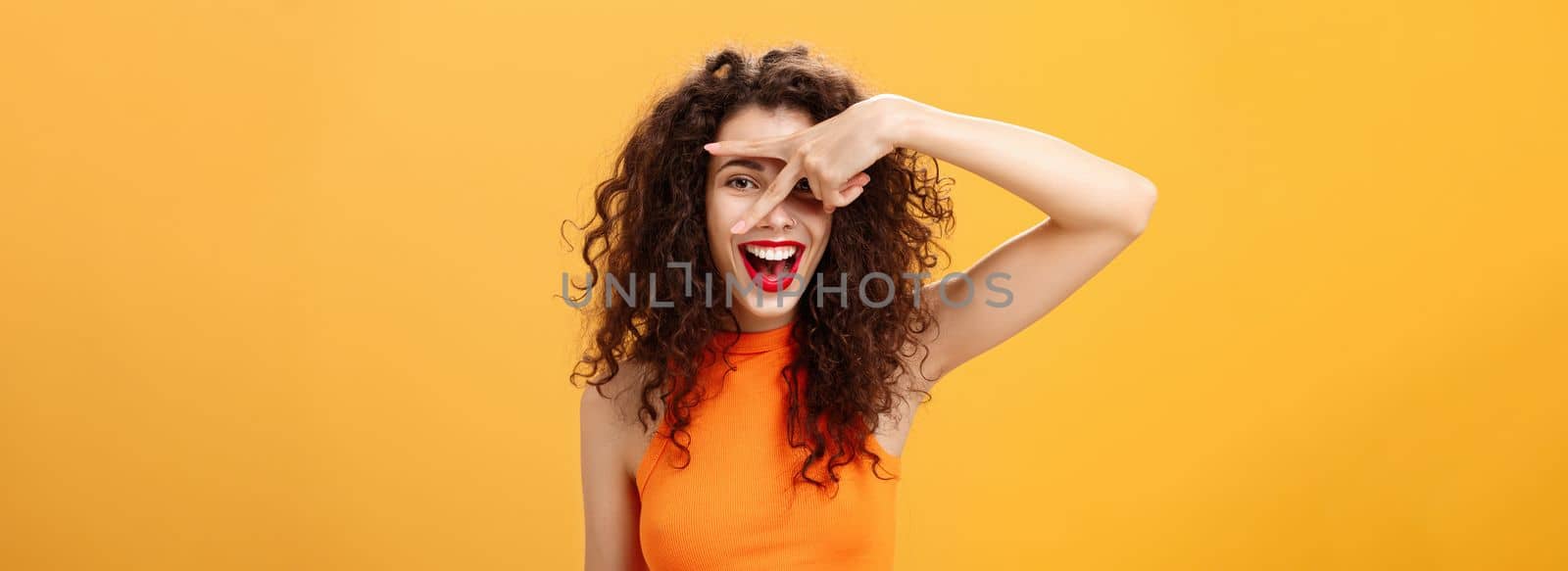 Waist-up shot of creative joyful caucasian female. with curly hairstyle and small tattoo showing peace gesture and peeking through fingers at camera happily smiling broadly having fun over orange wall.