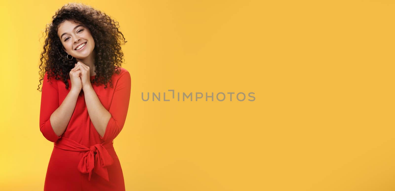 So sweet love you. Portrait of tender and silly cute curly-haired young woman in red dress tilting head holding hands together near face and smiling touched during heartwarming moment of confession.