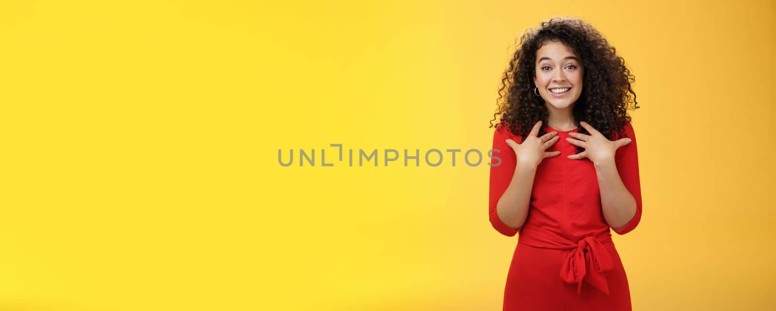 Pleased and thankful tender curly-haired kind girl in red dress pressing hands to breast being surprised with unexpected tender heartwarming gift thanking smiling delighted over yellow background by Benzoix