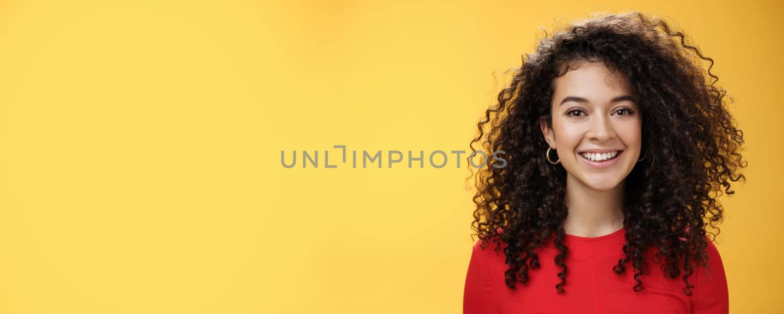 Close-up shot of pretty caucasian girl with curly hair in red dress and earrings smiling joyfully with pleased hopeful expression gazing at camera carefree, posing over yellow background.