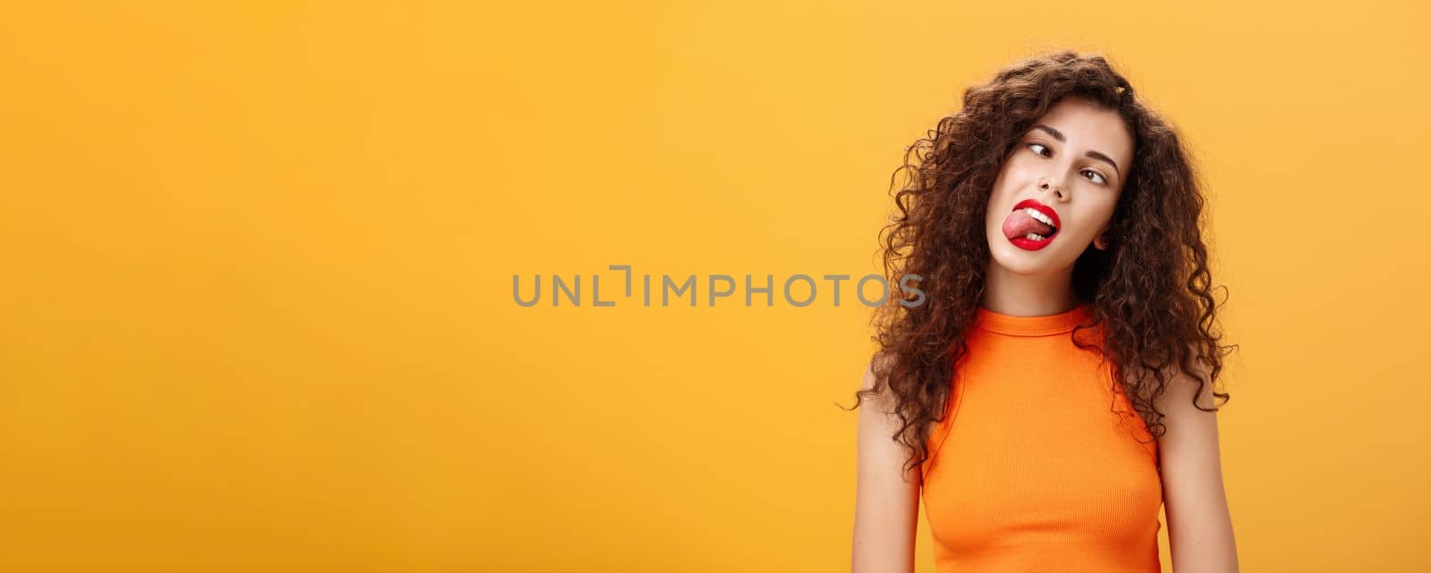 Girl fooling around carefree. feeling bored trying amuse herself during boring meeting sticking out tongue squinting and tilting head behaving childish and immature posing over orange background.