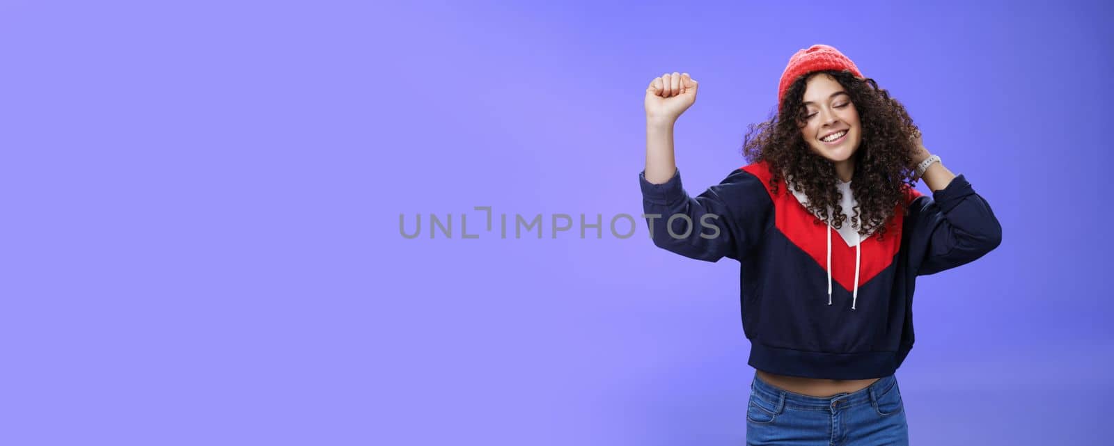 Girl delighted and carefree dancing over blue background with closed eyes and tender smile moving raised hands in rhythm of music, dreaming having positive mood wearing warm winter hat and sweatshirt.