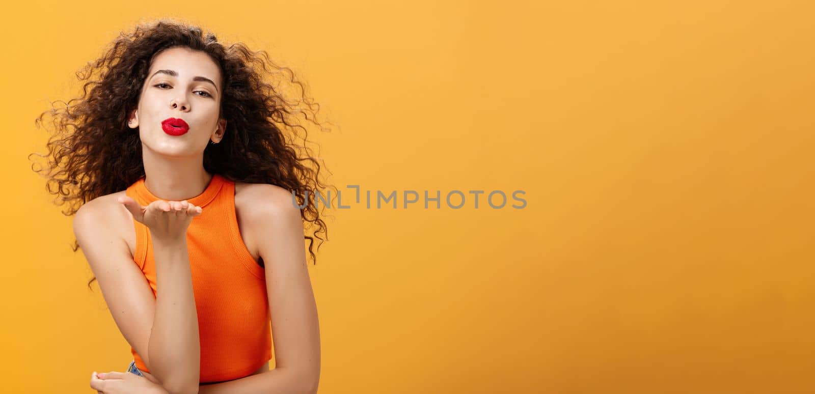 Flirty confident and sensual stylish woman with curly hairstyle and red lipstick folding lips holding palm near mouth sending kiss while hair flicks on air being in romantic mood over orange wall. Body language concept