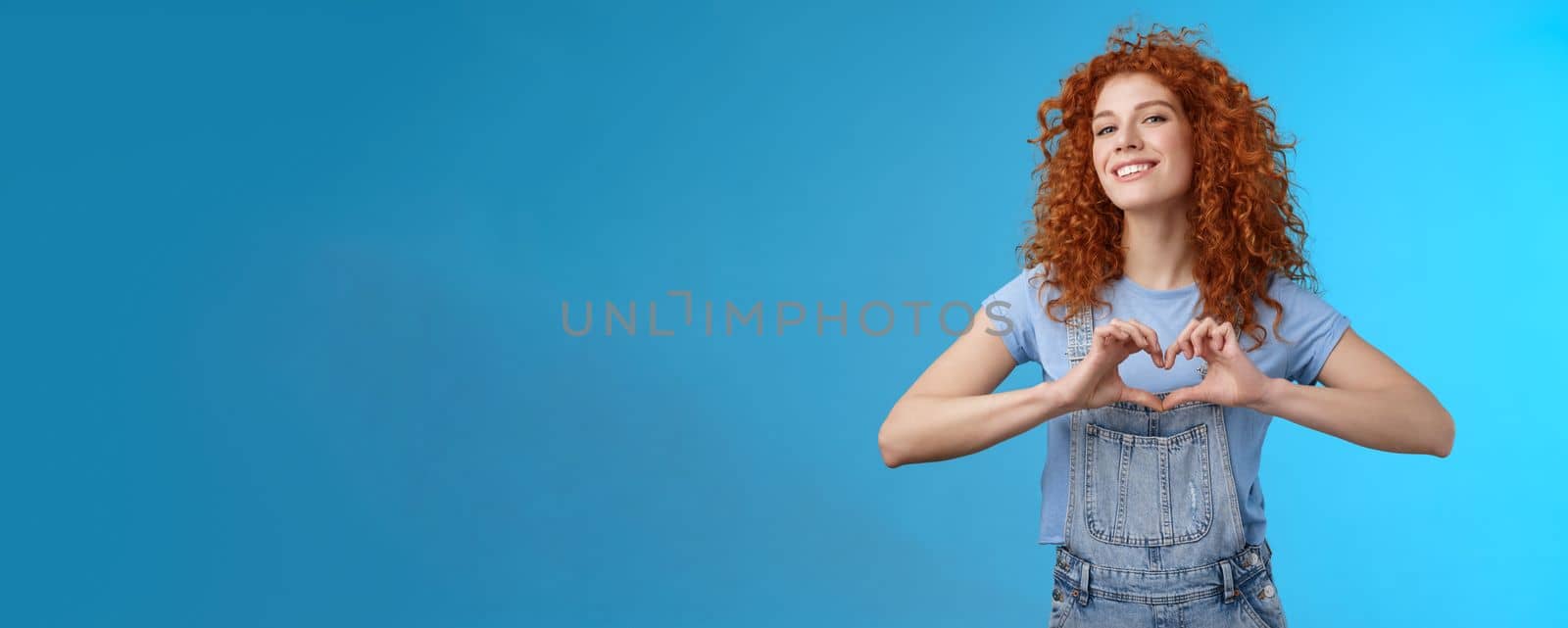 Sassy confident good-looking modern redhead curly woman raise head proud love tell girlfriend romantic heartwarming feelings present own heart smiling broadly standing blue background.