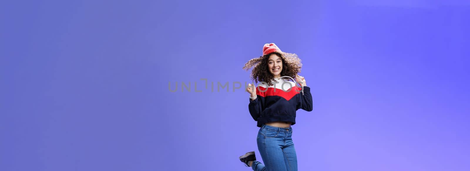Lifestyle. Delighted carefree and happy young woman with curly hair in warm beanie jumping joyfully having fun raising hands and smiling broadly posing over blue background in outdoor clothes.