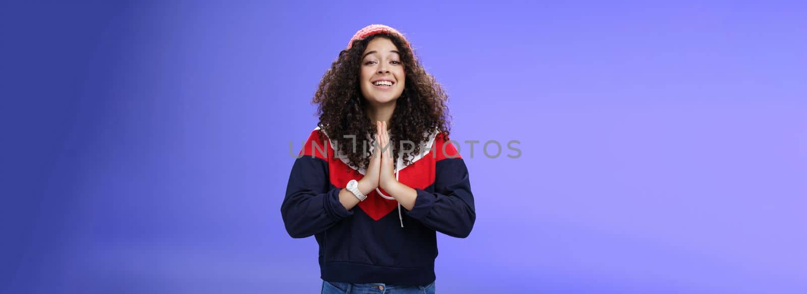 Lifestyle. Charming excited and friendly-looking cute female friend with curly hairstyle in outdoor hat and sweatshirt holding hands in pray and smiling with angel expression as hoping friend help over blue wall.