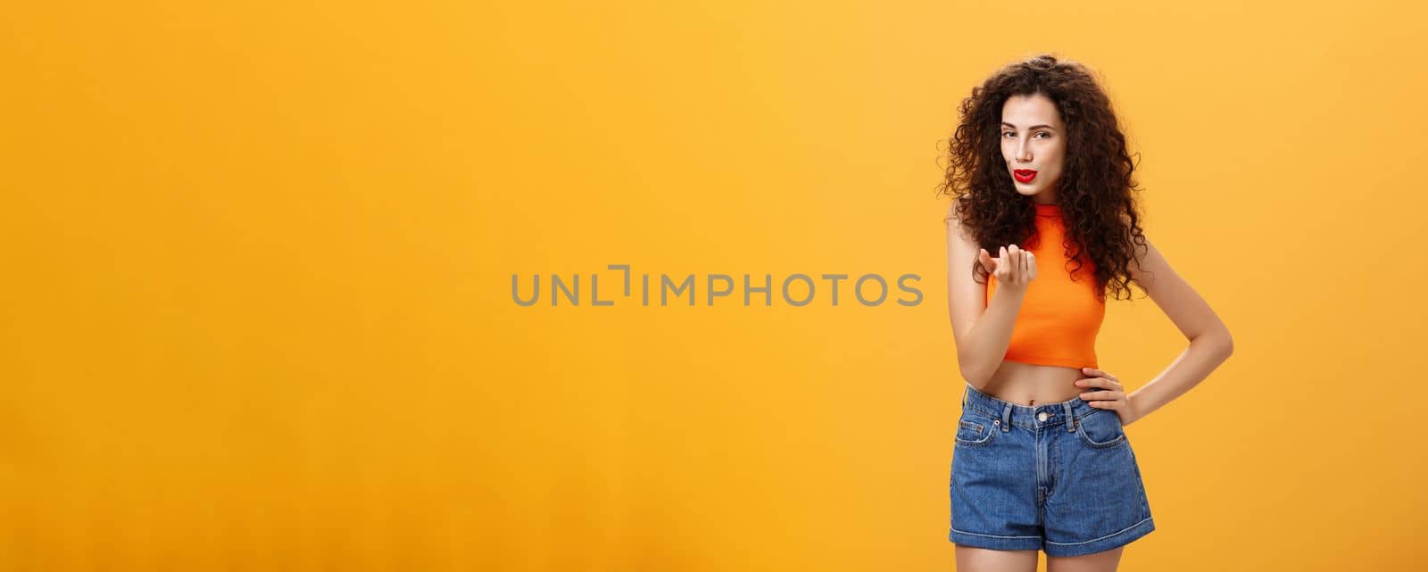 Woman seducing hot guy. Sensual and sexy daring female with curly hairstyle in stylish modern cropped top and shorts showing come here gesture inviting move closer flirting over orange background.