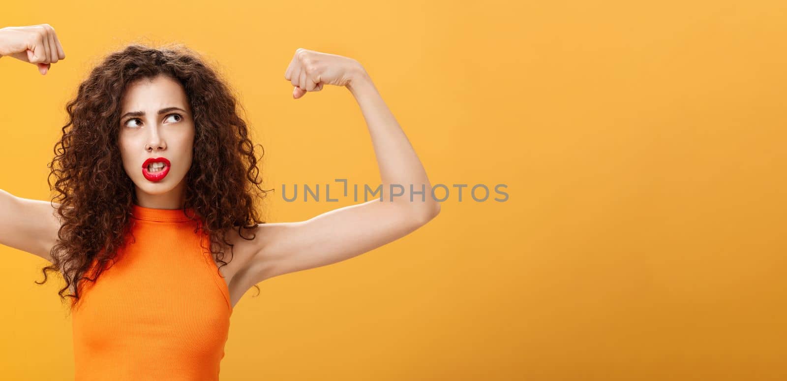Woman feeling powerful and strong raising hands. with clenched fists making intense face being working out in gym showing muscles and biceps looking at upper right corner posing over orange background.
