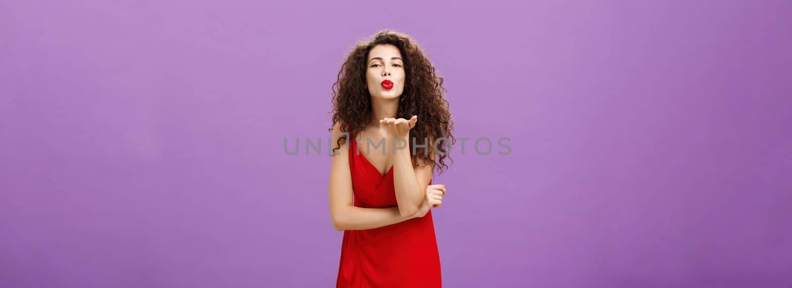 Woman sending passionate kiss being hopeless romantic. standing in elegant red dress with curly hairstyle and makeup blowing mwah with smile and folded lips holding palm near mouth over purple wall.