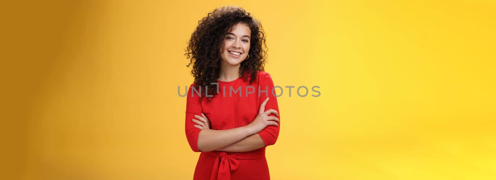 Confident charismatic young curly-haired woman in casual red dress holding hands crossed over chest in self-assured satisfied expression, smiling delighted posing over yellow background.