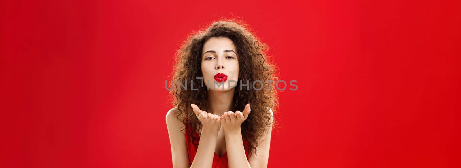 Woman rewards boyfriend with passionate wind kiss after romantic date. Portrait of sensual hot adult female in red elegant dress with curly hairstyle bending towards camera sending mwah flirty. Romance concept