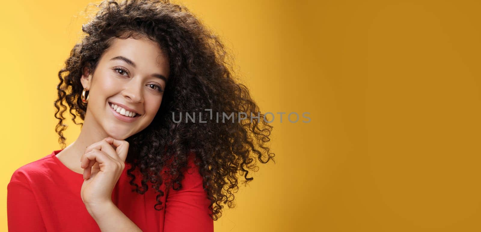 Close-up shot of stylish and happy bright curly-haired female in red dress tilting head sensually touching chin with finger and smiling broadly making flirty gazed at camera over yellow background by Benzoix