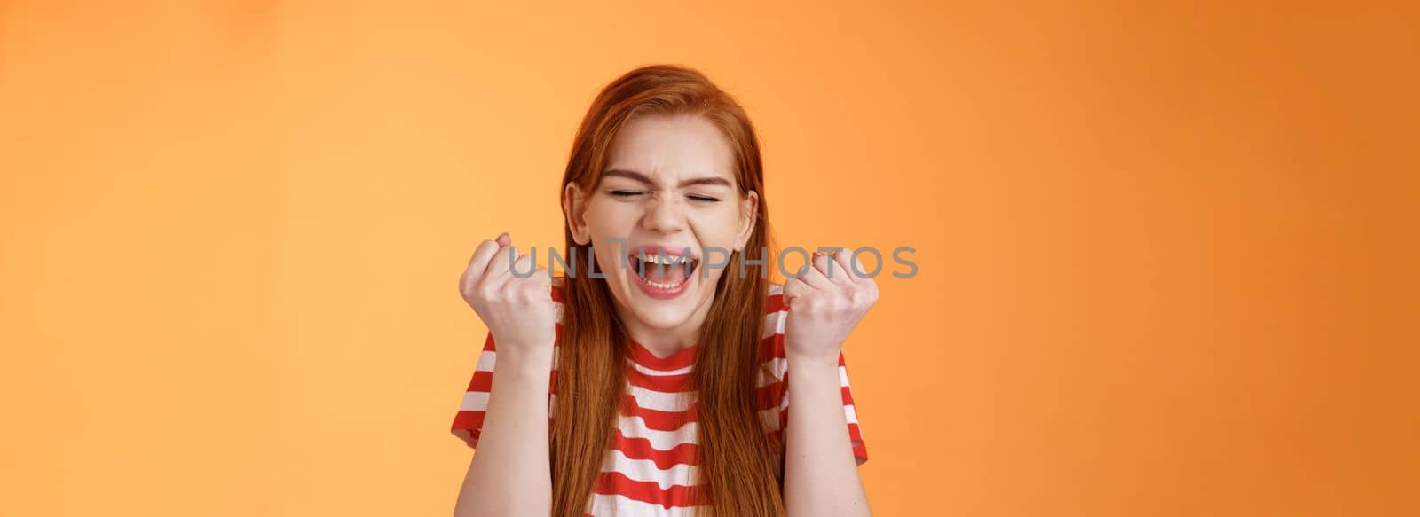 I did it. Charming lucky determined redhead female achieve success, fist pump celebrating, close eyes yell yes yeah, hooray gesture, triumphing victory, very good news, stand orange background.