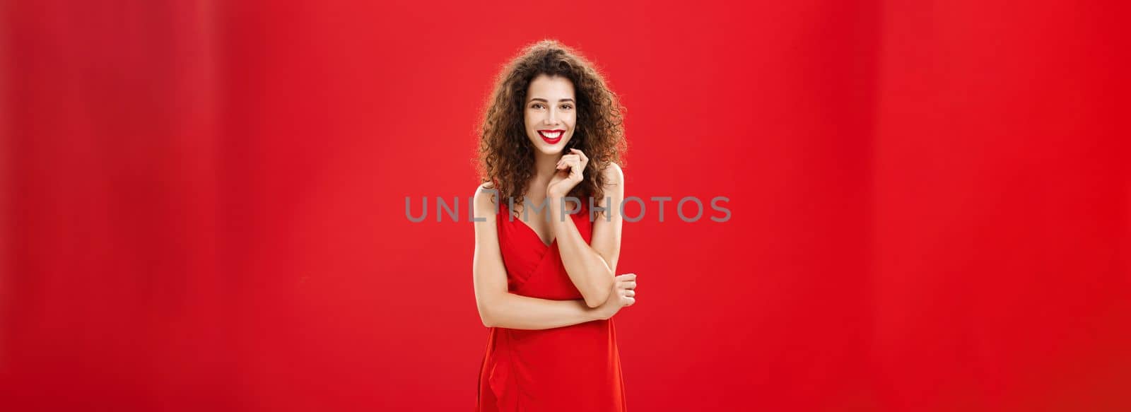 Passionate charming european woman over red background in elegant dress with curly hairstyle smiling cute, feminine playing with hair strand standing timid and silly, talking to person she admires.