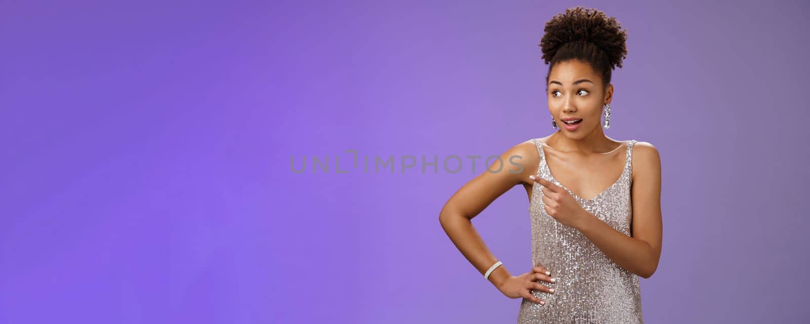 Curious unsure african-american attractive elegant young woman in silver glittering evening dress look pointing left intrigued discuss strange person gaze doubtful uncertain, blue background.
