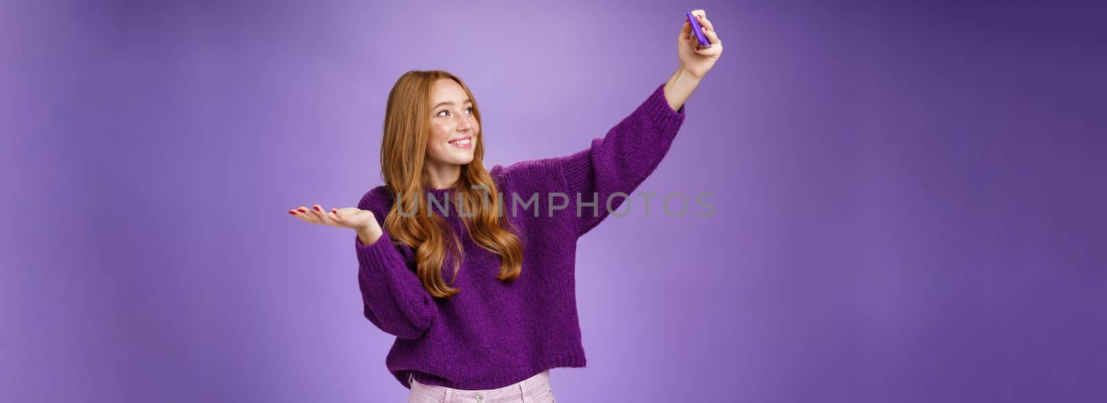 Portrait of cute redhead girl on vacation taking selfies near sightseeing raising hand as if holding builting on palm or showing cool place as taking photograph with pulled arm and smartphone. Technology and travel concept