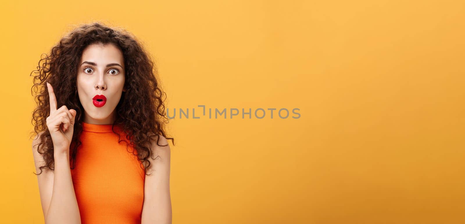 I got excellent plan. Enthusiastic emotive and excited female with curly hairstyle in orange cropped top raising index finger in eureka gesture folding lips popping eyes adding amazing suggestion.