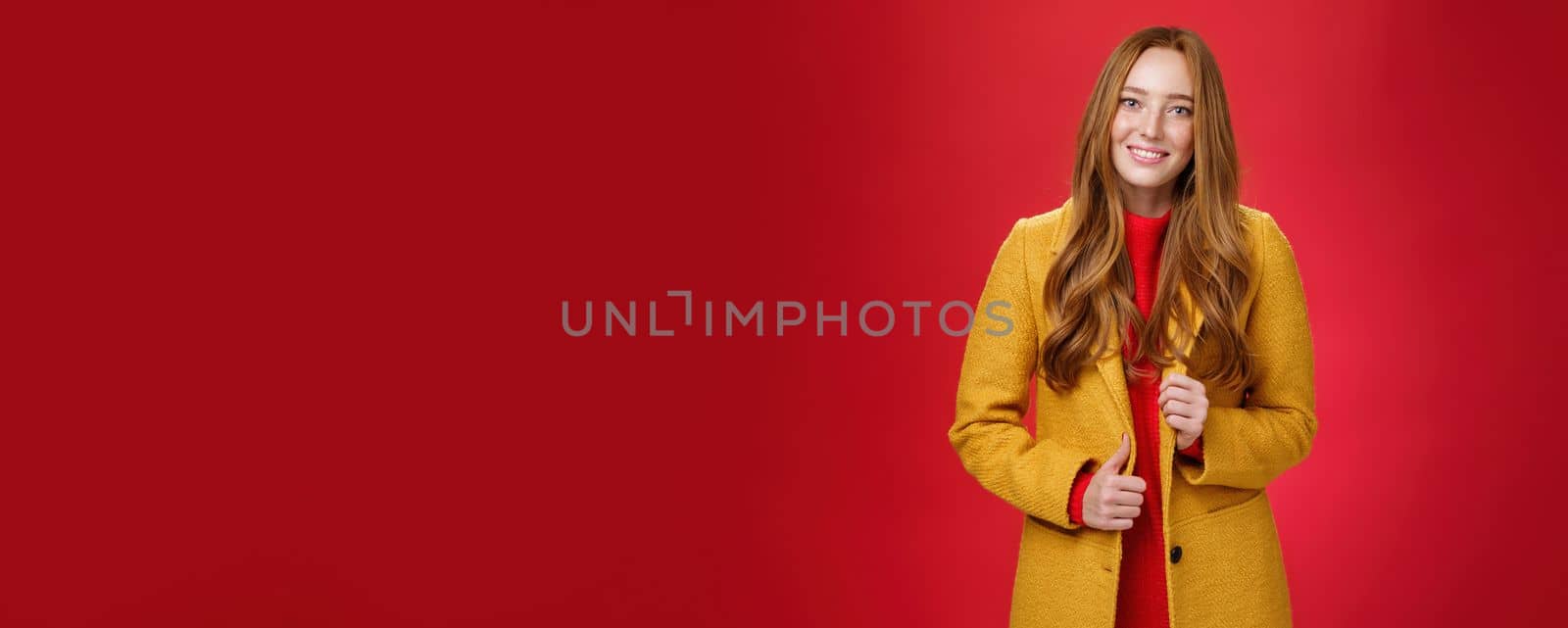 Stylish and cute good-looking redhead female in yellow coat on way to work, grabbing coffee making order with cute friendly smile touching button and posing over red background.