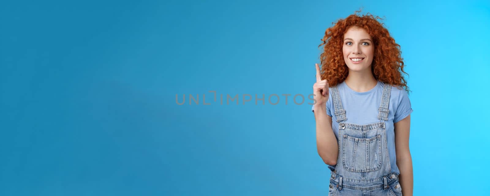 Cheerful cute energized redhead curly girl wear denim overalls book summer vacation trip offer good offer link pointing raised index finger up show uppwards advertisement smiling silly by Benzoix