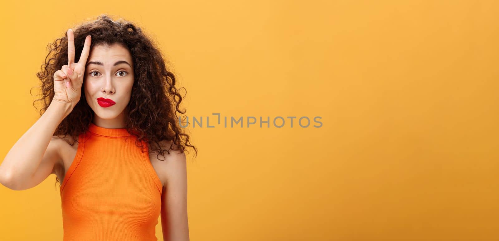 Waist-up shot of charismatic good-looking european female with curly haircut in cropped top showing peace sign near face making silly look smirking and gazing friendly at camera over orange background by Benzoix