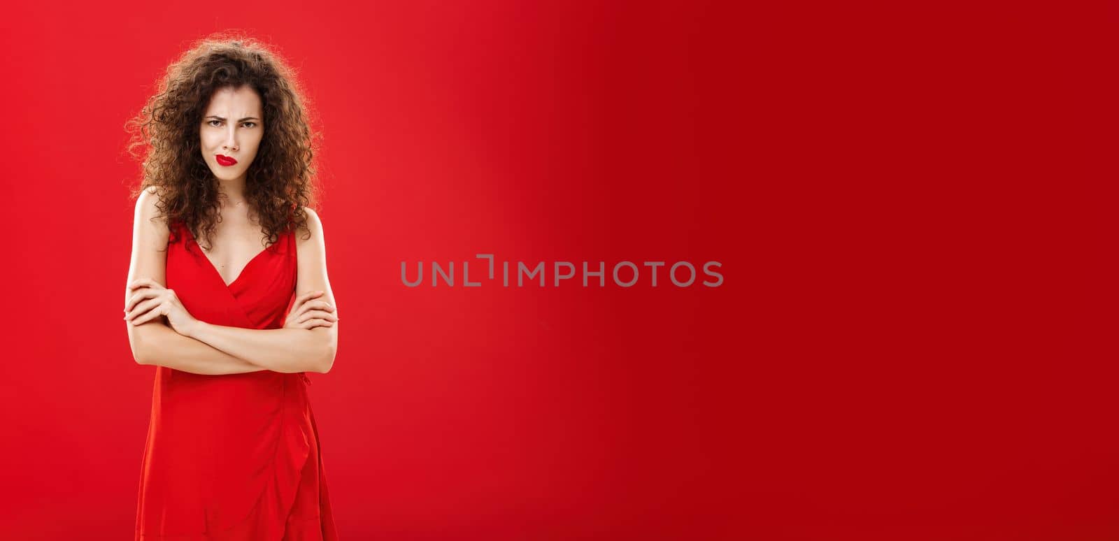 Portrait of intense thoughtful moody woman with curly hairstyle. frowning smirking looking from under forehead crossing arms against chest in offended and defensive pose over red background.