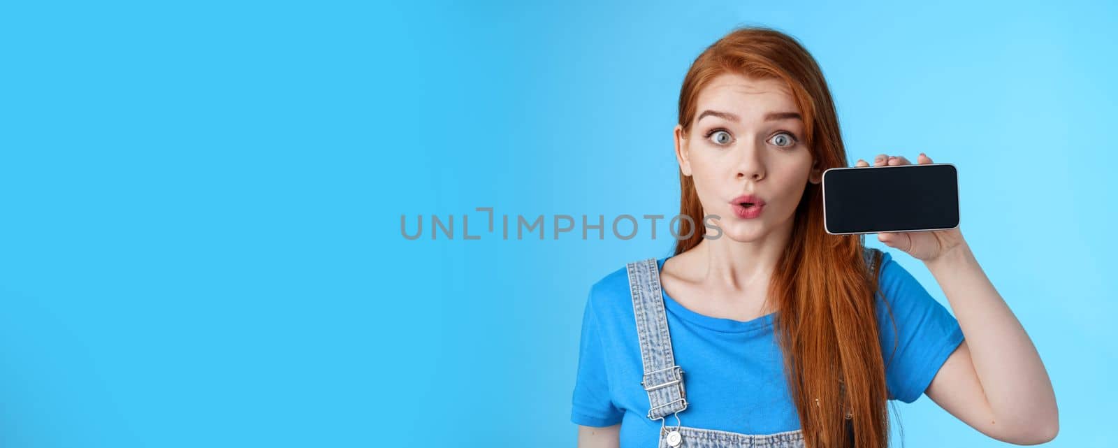 Amazed fascinated excited 20s redhead woman show smartphone screen horizontally, fold lips whistling amused wondered, check out cool phone app, stand blue background curious. Copy space