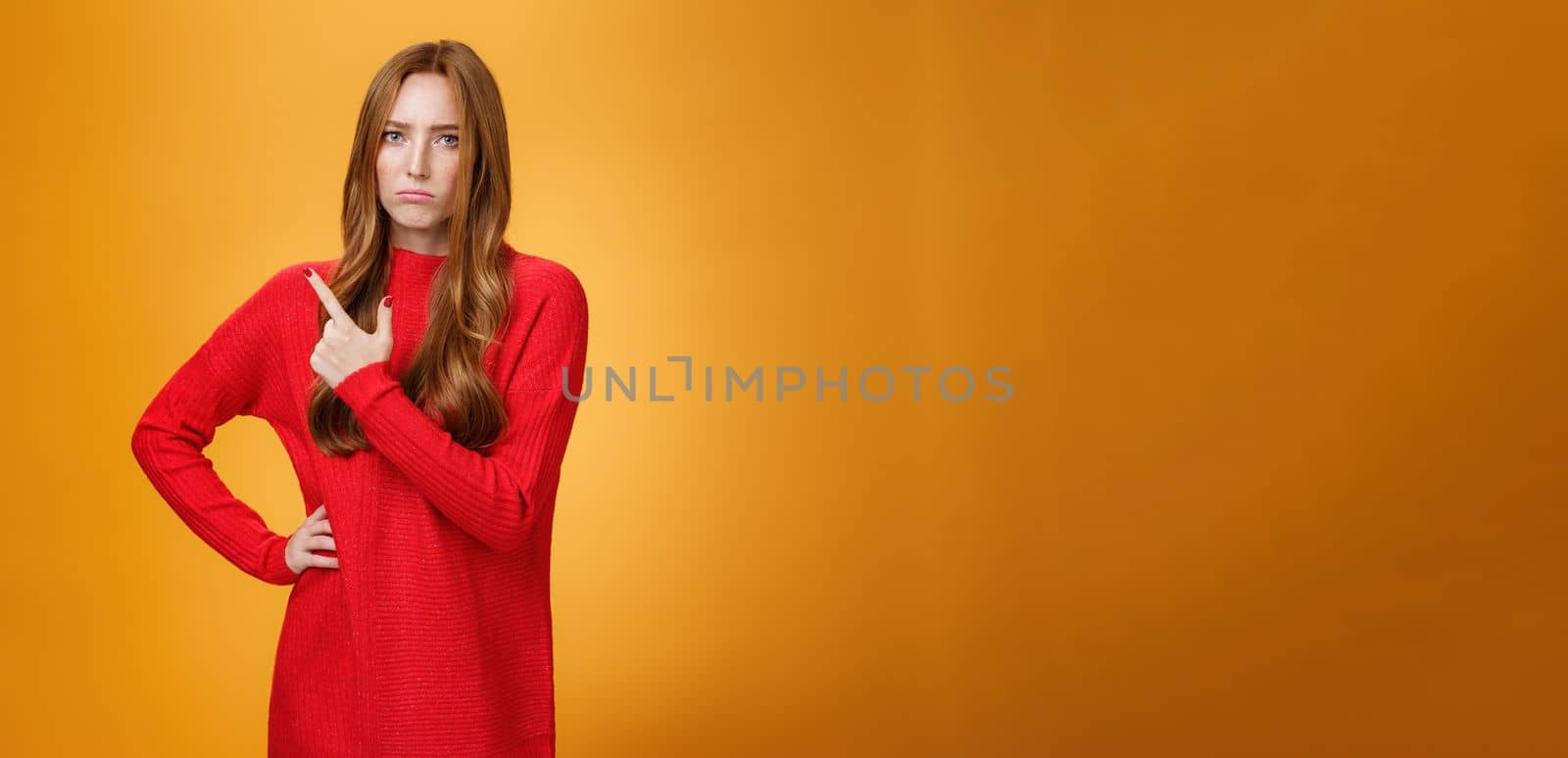 Upset and disappointed young ginger girl in red sweater pointing at upper left corner or behind frowning looking gloomy and angry, standing jealous and moody against orange background.