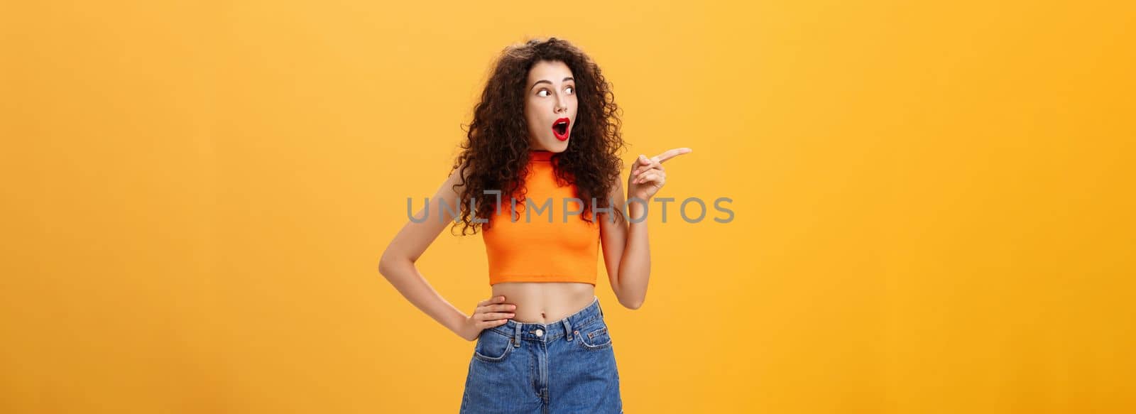 Woman peeking at neighbours window. being surprised and amazed pointing and staring speechless left standing over orange background with dropped jaw intrigued and thrilled look in cropped top.