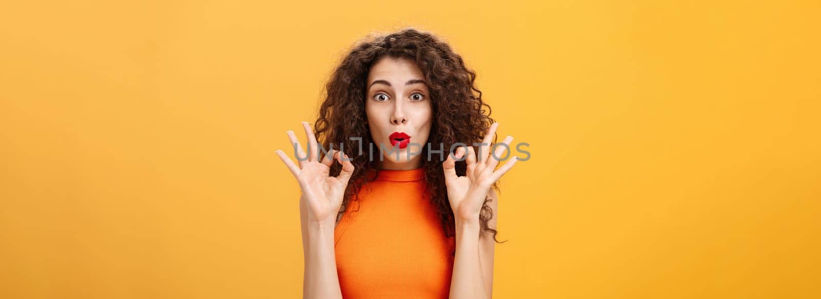 Enthusiastic charming caucasian girl. with curly hairstyle in red lipstick and orange top showing okay gesture being intrigued and delighted folding lips approving and agreeing on awesome adventure.