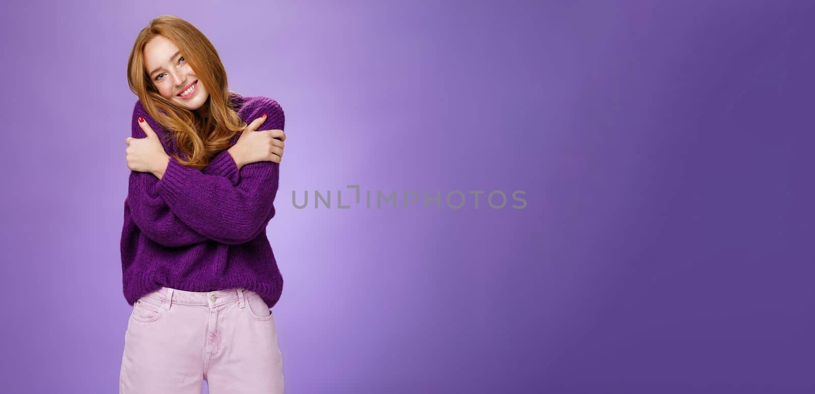 Lifestyle. Silly and cute carefree redhead woman in 20s leaning on shoulder as hugging herself feeling warmth wearing purple sweater smiling broadly in cozy and relaxing atmosphere over violet wall.