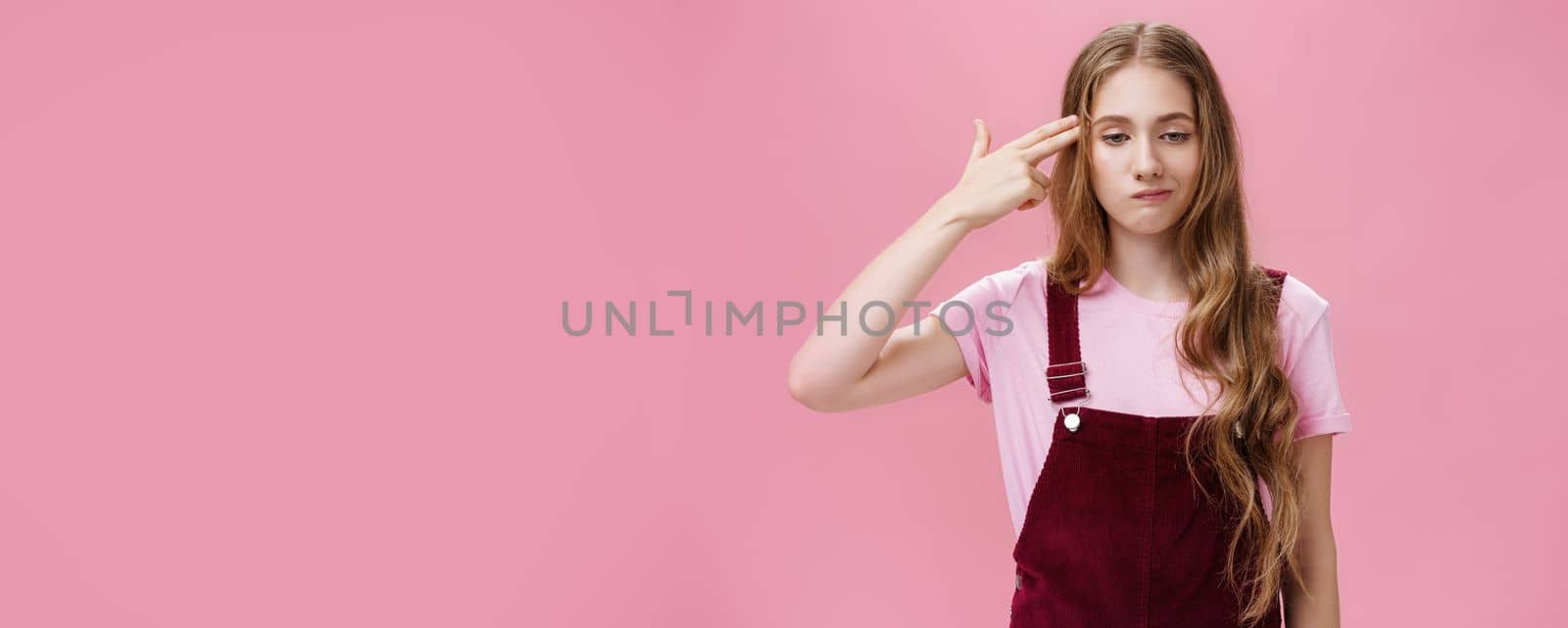 Portrait of bothered uneasy cute european female with long natural fair hair in trendy corduroy overalls smirking from irritation and annoyance showing finger fun gesture as if shooting in head. Copy space