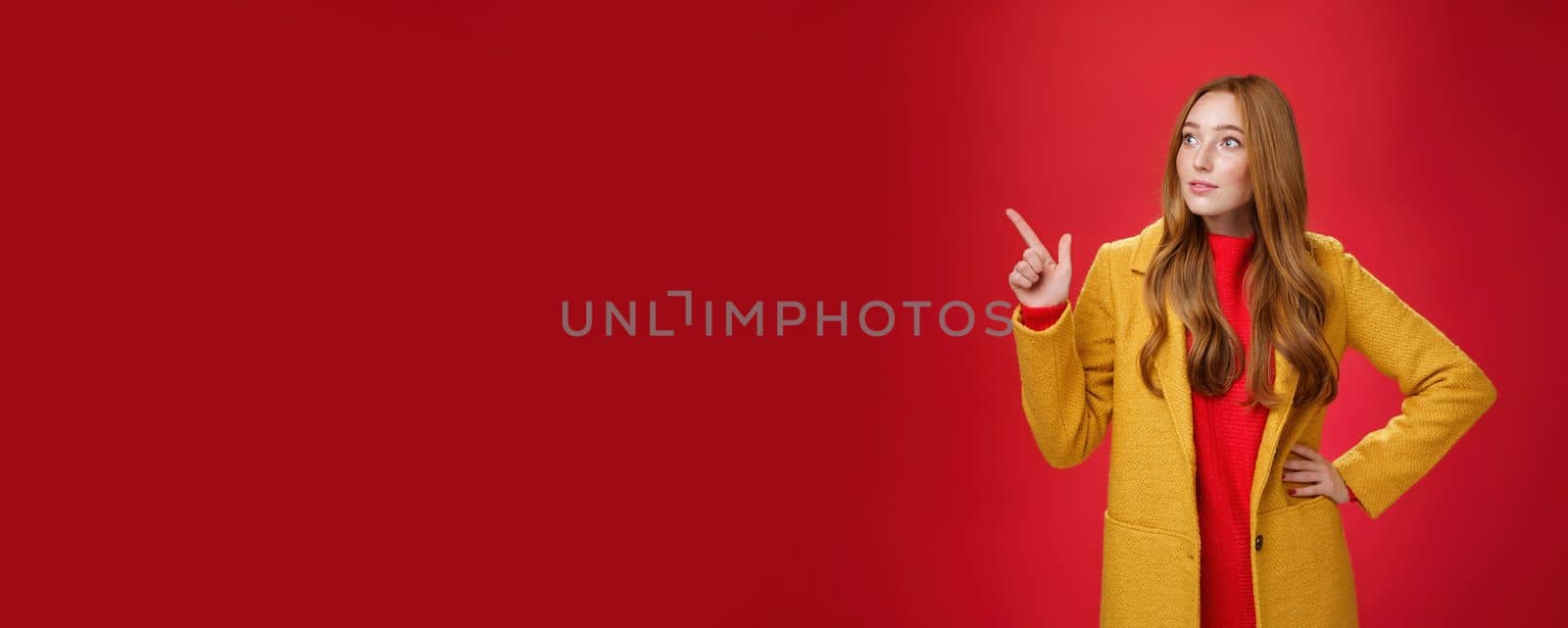 Wondered curious good-looking redhead woman in coat stopping on way looking and pointing at upper left corner curious and interested, observing surprising promotion over red background.
