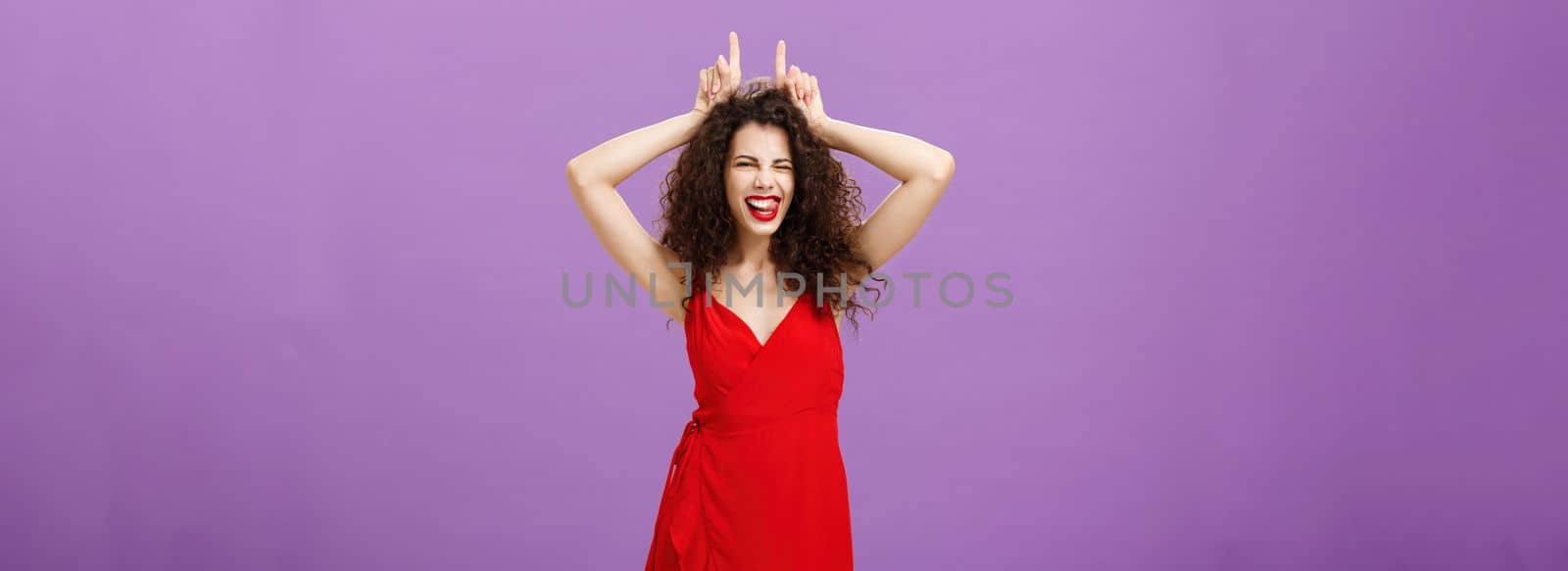 Devil lives inside lady. Daring stylish adult woman with curly hairstyle in red evening dress winking making confident and amused expression showing horns with index fingers on head, being stubborn.