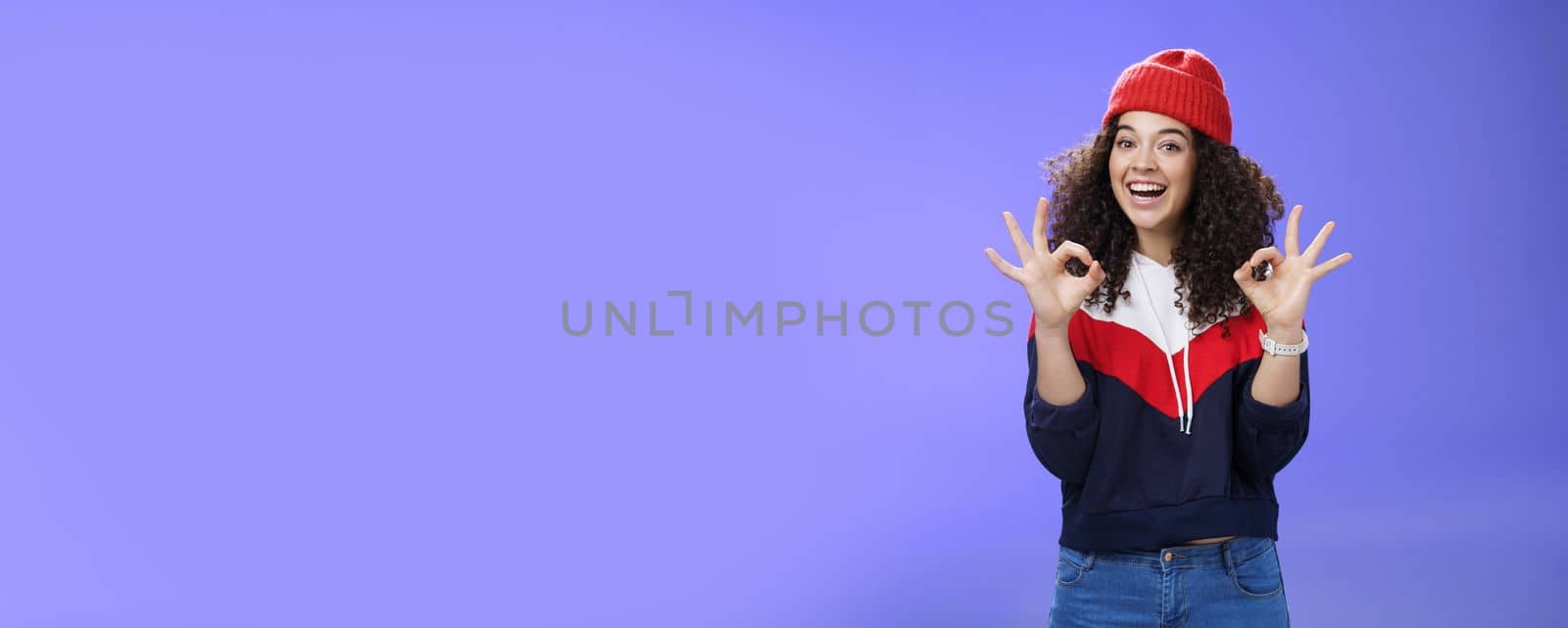 Got under control. Portrait of happy charming smiling curly-haired female in warm winter hat and sweatshirt smiling broadly and showing okay or excellent gesture as approving, liking cool movements by Benzoix